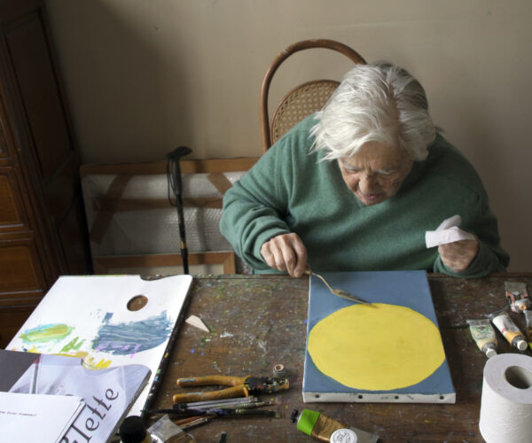 Etel Adnan sitting at table painting surrounded by painting supplies