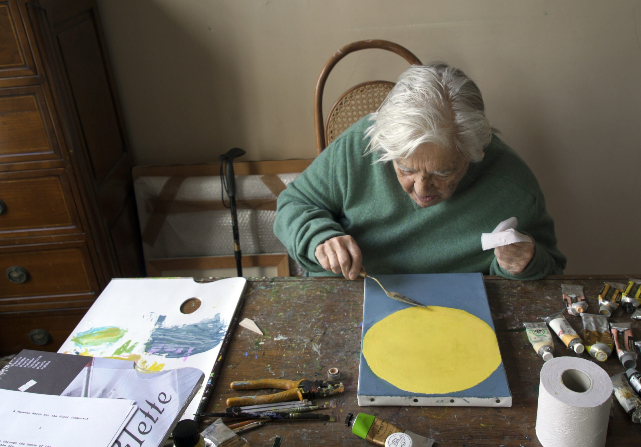 Etel Adnan sitting at table painting surrounded by painting supplies